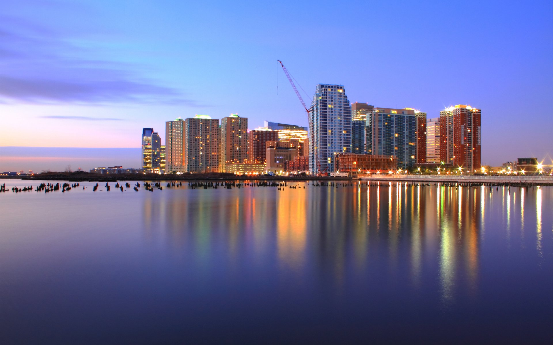 HD desktop wallpaper showcasing the illuminated Jersey City skyline at dusk, reflecting on calm waters under a clear blue sky.