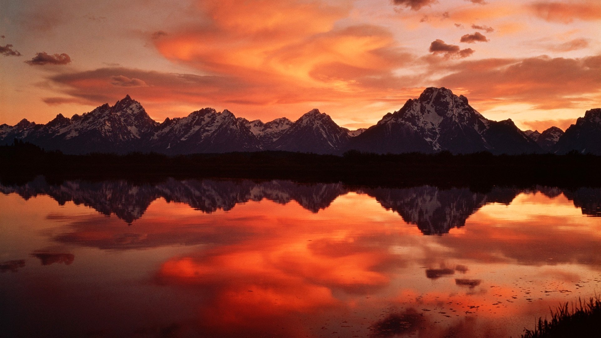 HD PC desktop wallpaper showcasing a vibrant orange sunset reflecting perfectly on a calm lake with silhouetted mountain peaks in the background.