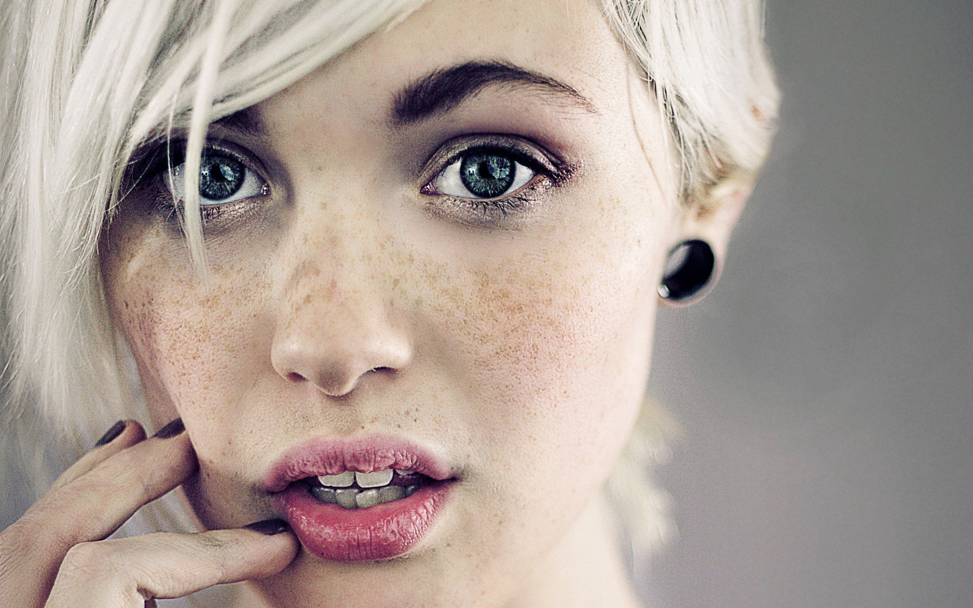 A close-up of a woman with short, platinum blonde hair and striking green eyes, featuring freckles and subtle makeup, set against a soft, neutral background. High-definition wallpaper for desktop use.