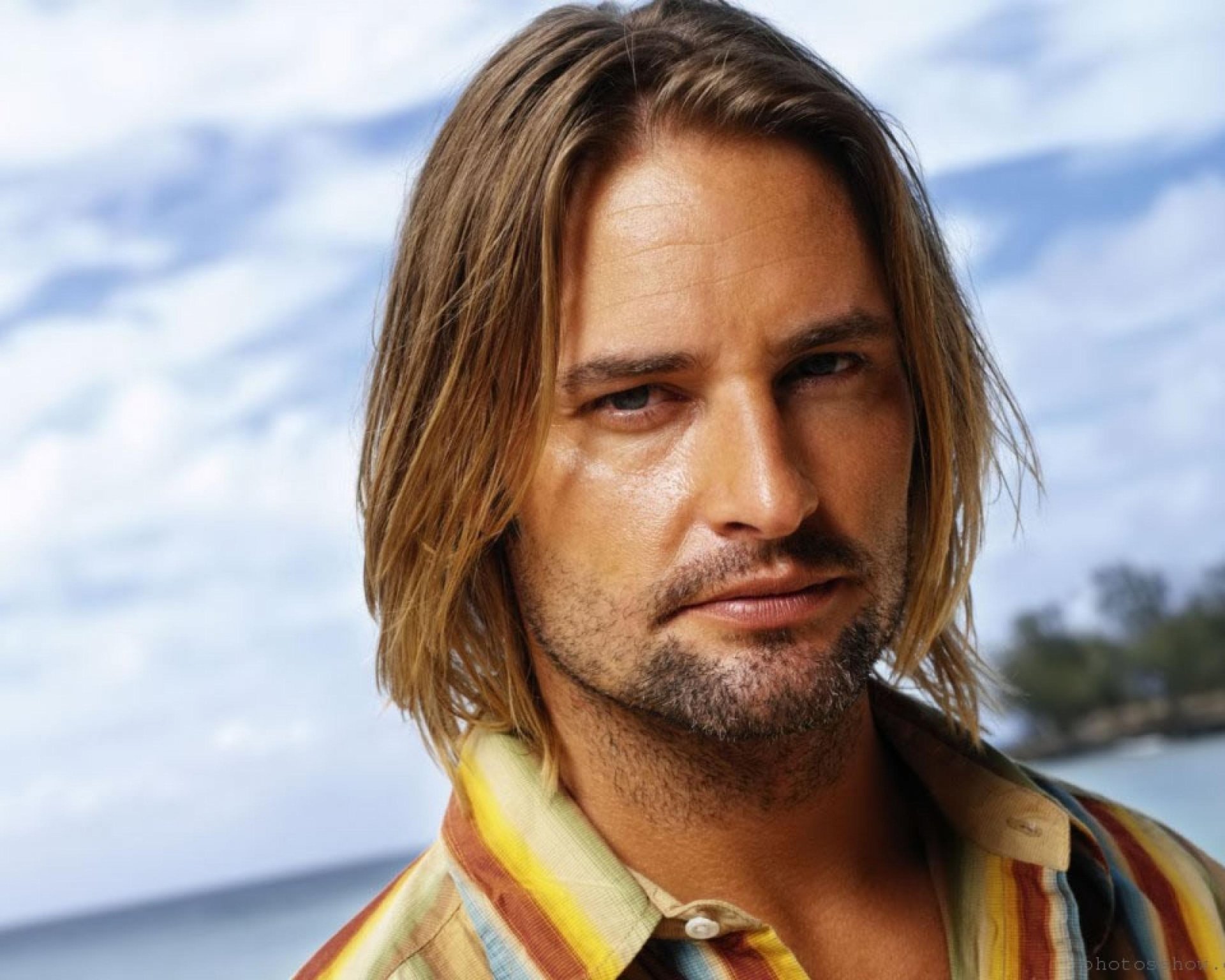 HD desktop wallpaper featuring celebrity Josh Holloway with shoulder-length hair, set against a bright, partly cloudy sky background.