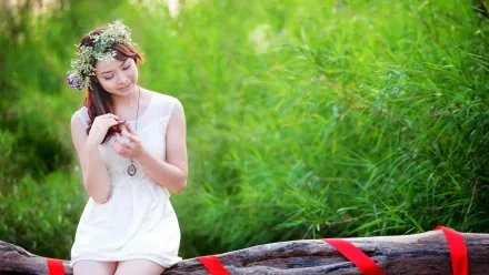 A beautiful woman in a white dress sits on a log adorned with red ribbons, surrounded by lush greenery, wearing a flower crown. This high-definition image makes a stunning desktop wallpaper and background.