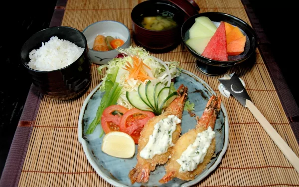 HD PC desktop wallpaper featuring a meal with fried shrimp topped with sauce, fresh salad, rice, miso soup, pickles, and fruit on a bamboo mat background.