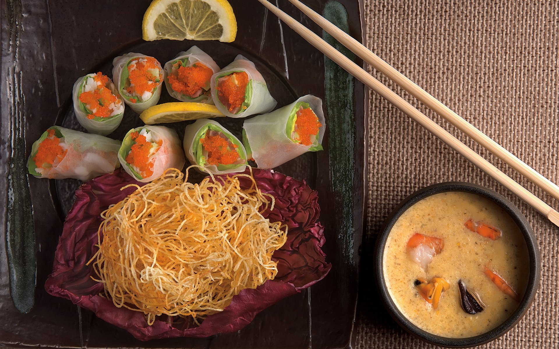 HD PC desktop wallpaper featuring a vibrant meal of fresh vegetable spring rolls, crispy noodles, and a bowl of seafood chowder with chopsticks on a rustic table.