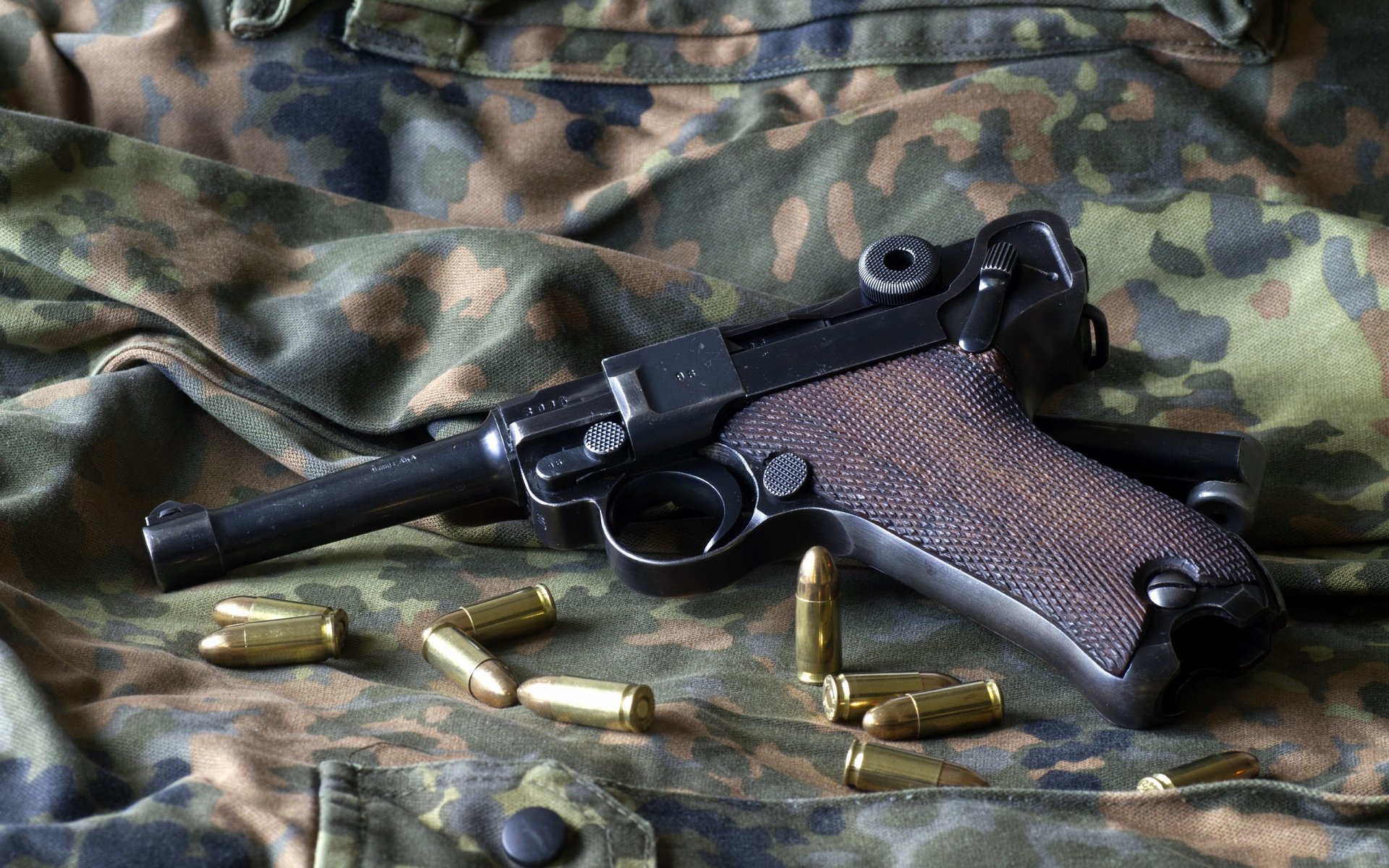 HD PC desktop wallpaper featuring a man-made Luger pistol resting on camouflage fabric with scattered brass bullet casings.