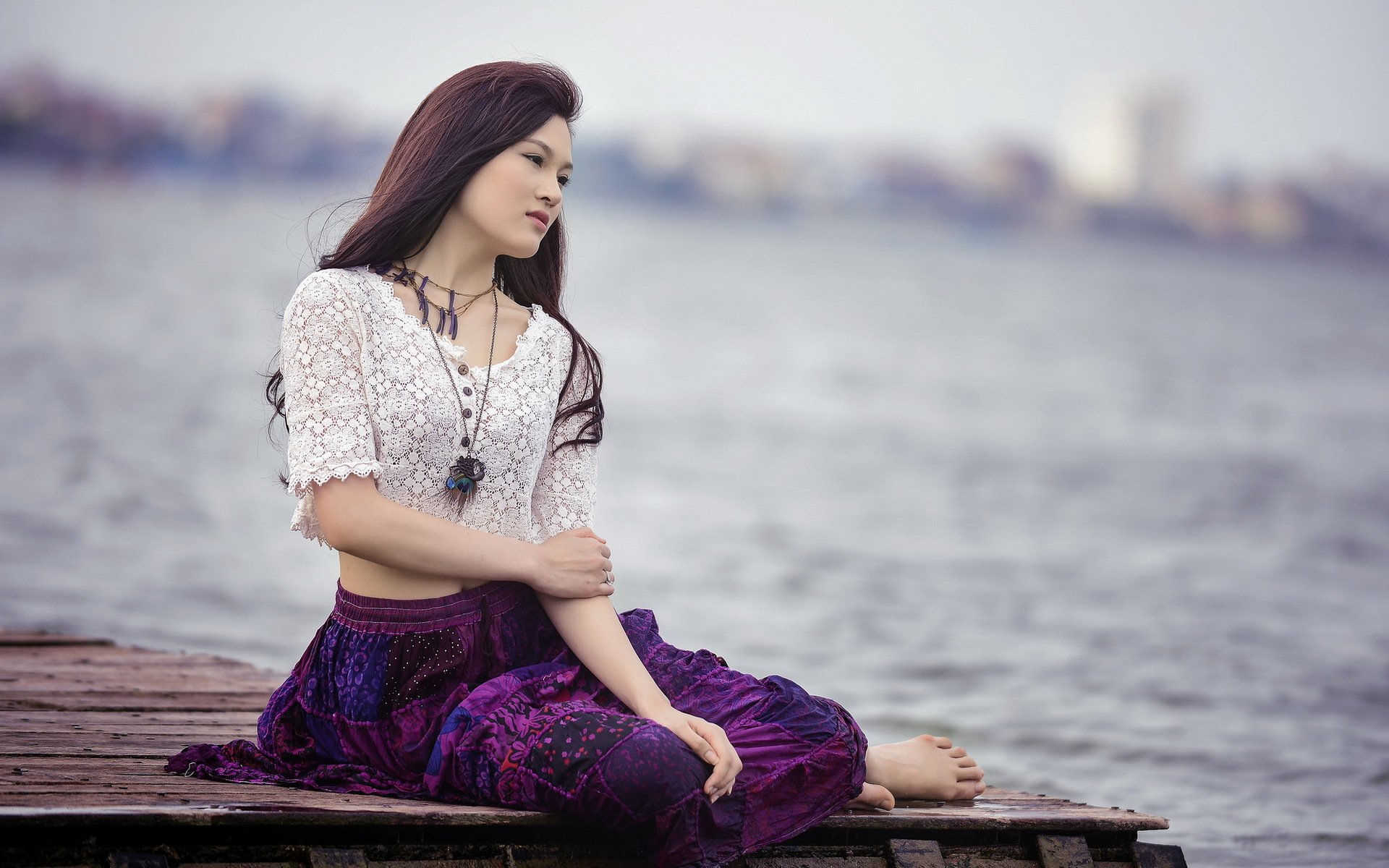 HD desktop wallpaper of an Asian woman sitting on a wooden dock by the water, gazing thoughtfully into the distance, dressed in a white blouse and purple skirt.