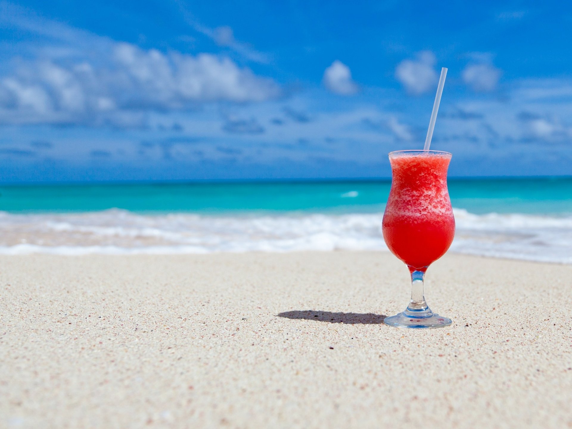 HD PC desktop wallpaper featuring a vibrant red tropical drink with a straw on a sandy beach, set against a bright blue sky and ocean backdrop.