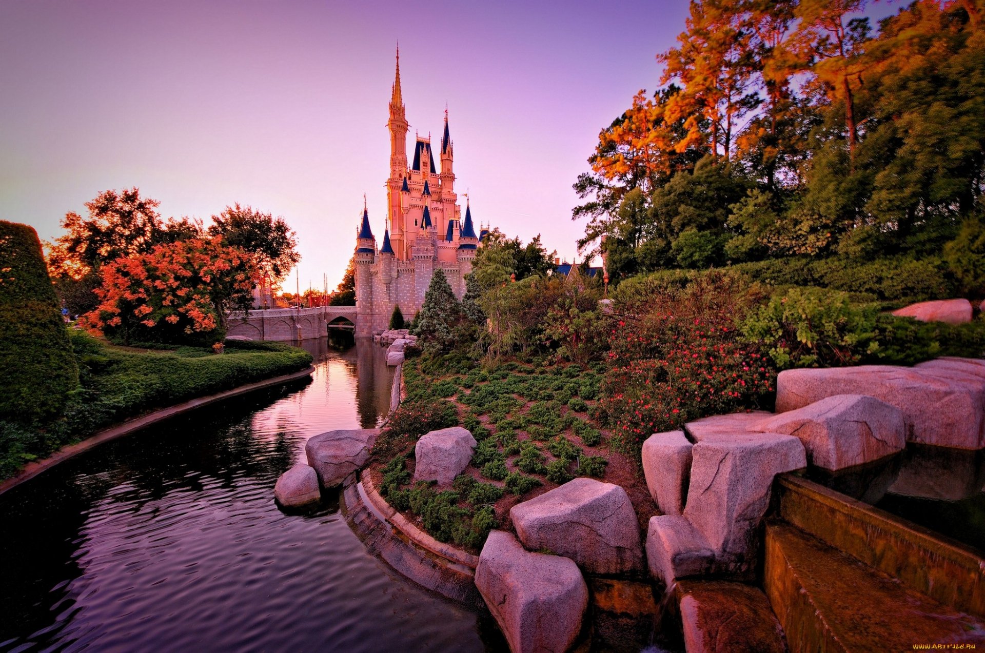 A stunning HD desktop wallpaper featuring a majestic castle surrounded by lush greenery and a serene waterway, illuminated by the warm glow of sunset hues.