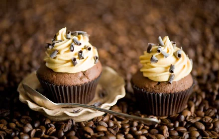 Two delicious cupcakes with creamy frosting and chocolate sprinkles sit on a plate, surrounded by scattered coffee beans, creating a warm and inviting food-themed HD wallpaper.