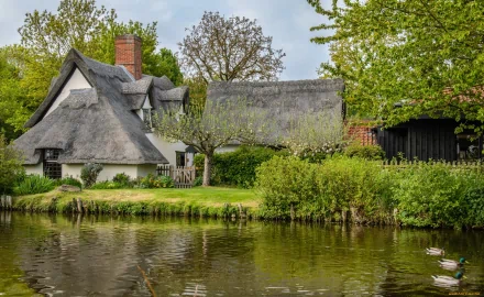A charming thatched-roof house surrounded by lush greenery, reflecting beautifully in the calm water nearby, makes an inviting HD desktop wallpaper and background.