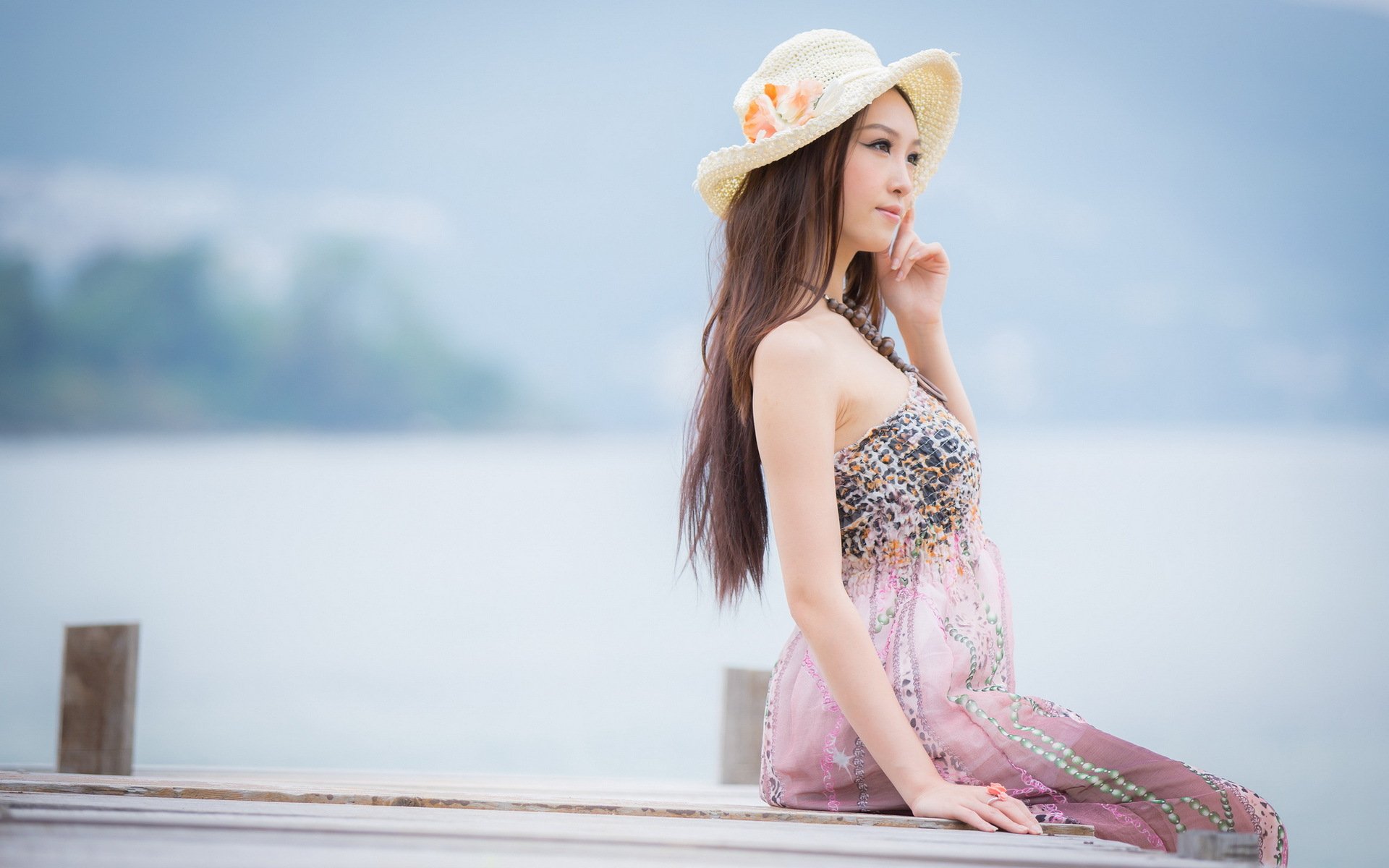 HD desktop wallpaper featuring an Asian woman in a summer dress and hat, sitting by a serene waterfront with a distant, blurred background.