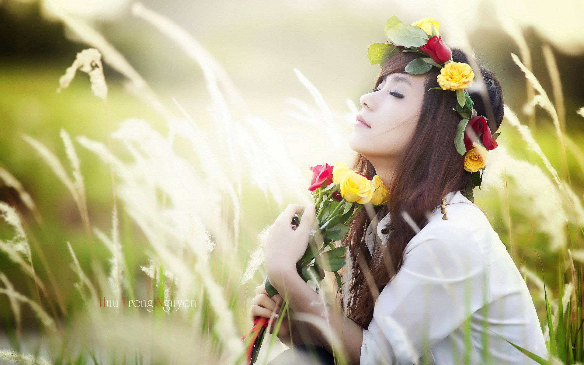 A serene Asian woman wearing a floral crown holds roses in a sunlit field, creating a tranquil and vibrant desktop wallpaper.