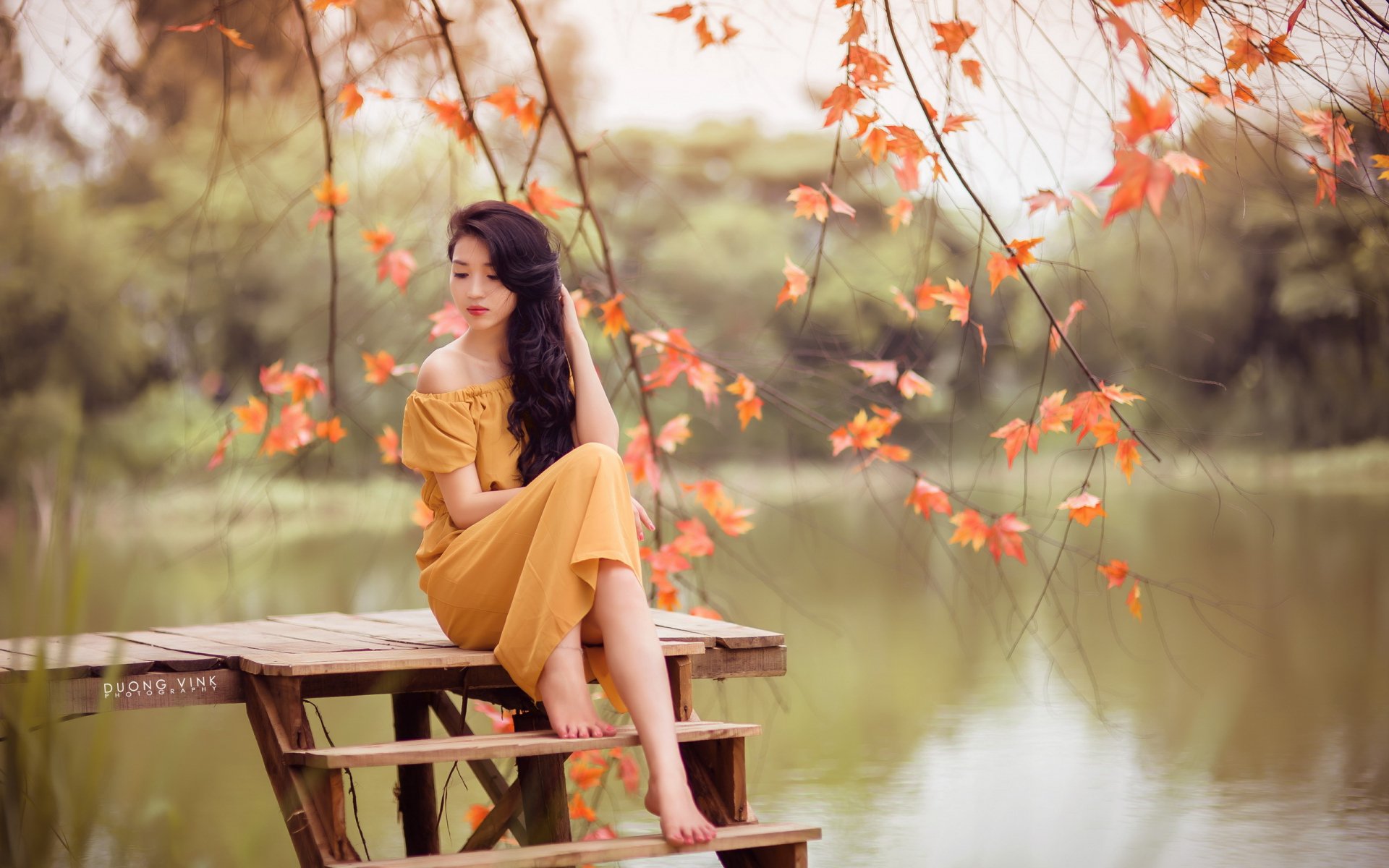 An Asian woman with long black hair and a yellow dress sits on wooden steps near a serene lake, surrounded by autumn leaves. The image has a shallow depth of field, adding a dreamy mood.