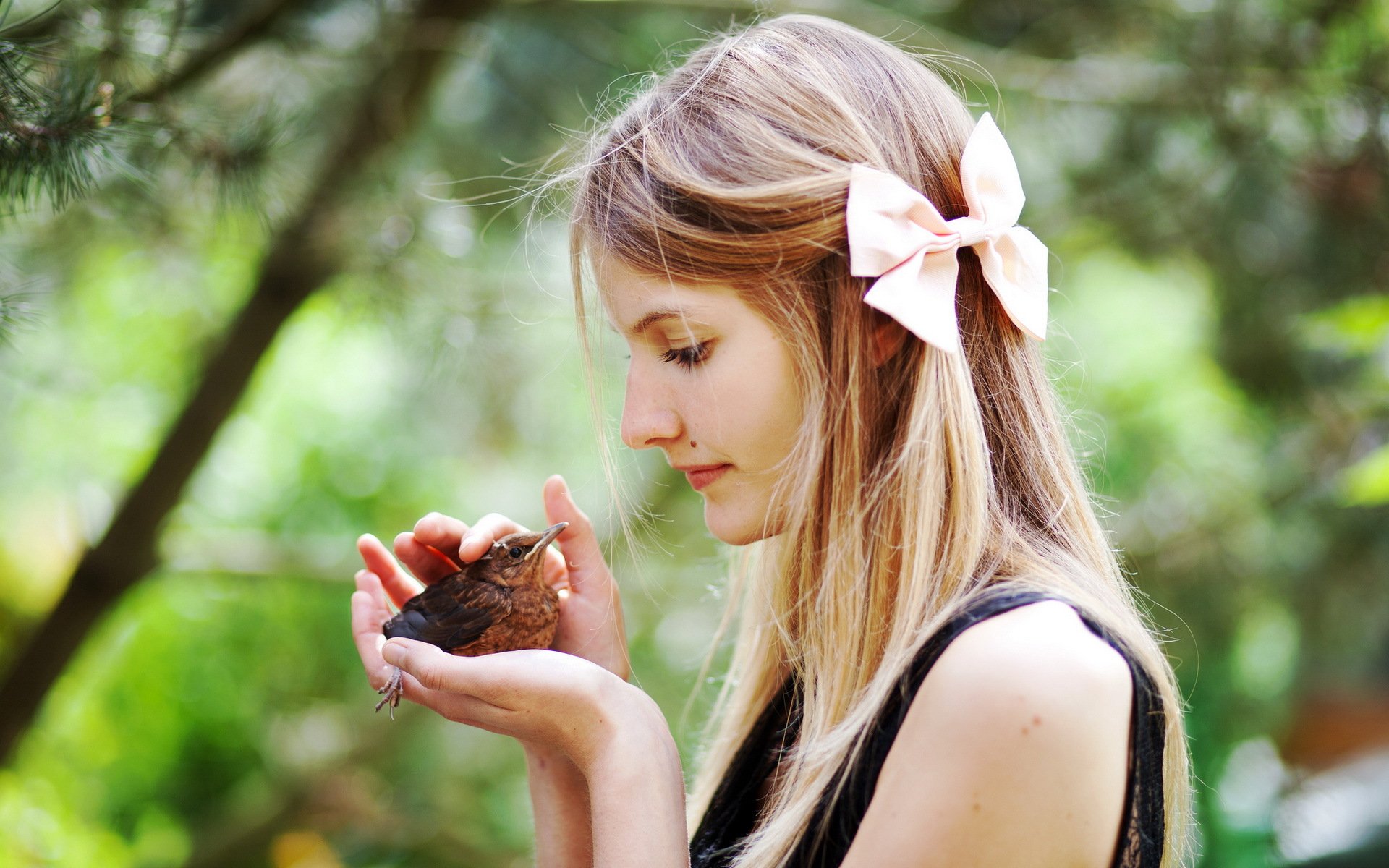 A serene image of a woman gently holding a small bird, adorned with a pink bow in her hair, surrounded by a lush, green backdrop, creating a calming atmosphere for desktop wallpaper.