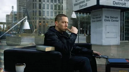 A contemplative Eminem sits on a sofa in an urban setting, reflecting on his music journey amidst the backdrop of Detroit. A stylish HD desktop wallpaper for fans.