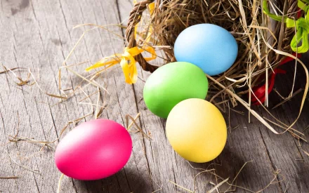 A vibrant Easter-themed desktop wallpaper featuring colorful eggs—pink, green, blue, and yellow—nestled in straw against a rustic wooden background.