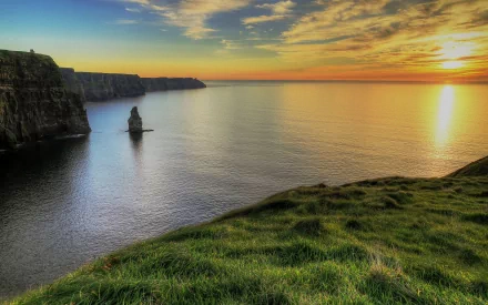 Sunset over the Cliffs of Moher coastline, with calm ocean waters and lush green grass in this HD PC desktop wallpaper.
