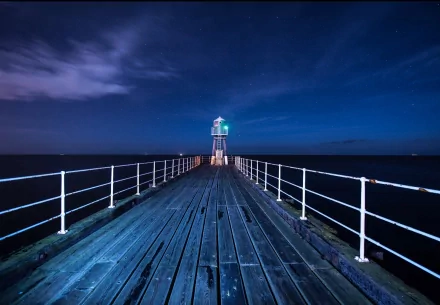 A serene view of a pier leading to a lighthouse at night, illuminated by a green light, showcasing man-made beauty against a tranquil sea and starry sky.