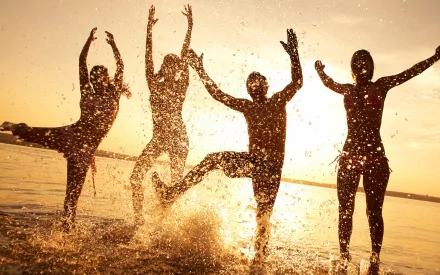 Four joyful people splash in the water against a vibrant sunset, capturing a moment of fun and laughter. This HD photography makes an engaging desktop wallpaper.