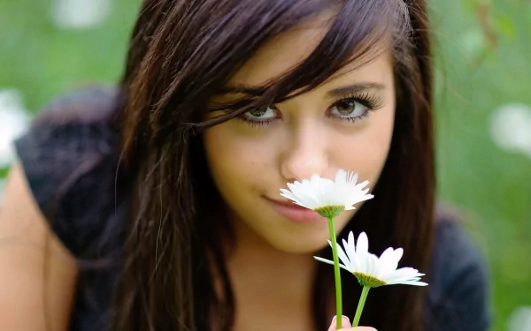 A woman with dark hair holds a white daisy close to her face, gazing intently at the camera. The background is a soft focus of green, creating an HD desktop wallpaper.