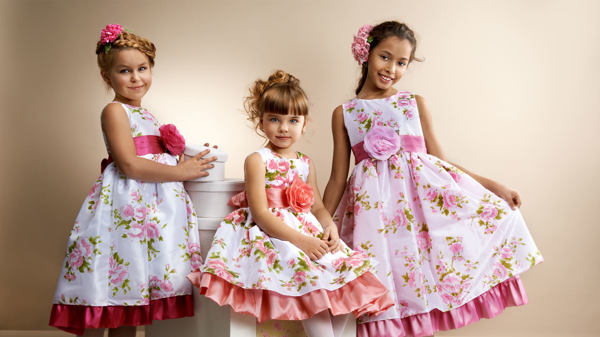 Three girls in stylish floral dresses pose together, showcasing their playful expressions against a soft background. This charming photography makes a delightful HD desktop wallpaper.