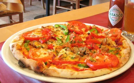 HD PC desktop wallpaper showing a fresh pizza topped with tomatoes, green peppers, and sausage, set on a table with a bottle of Tabasco sauce in the background.