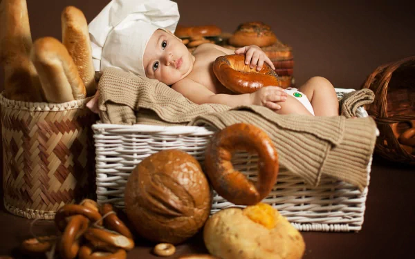 A baby wearing a chef’s hat lies in a wicker basket surrounded by various breads, captured in a 4K Ultra HD photography wallpaper.