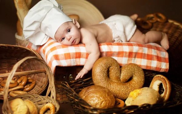 A charming baby wearing a chef's hat rests on a checkered cushion, surrounded by baskets of fresh bread and pastries, creating a delightful kitchen scene. 4K Ultra HD desktop wallpaper.