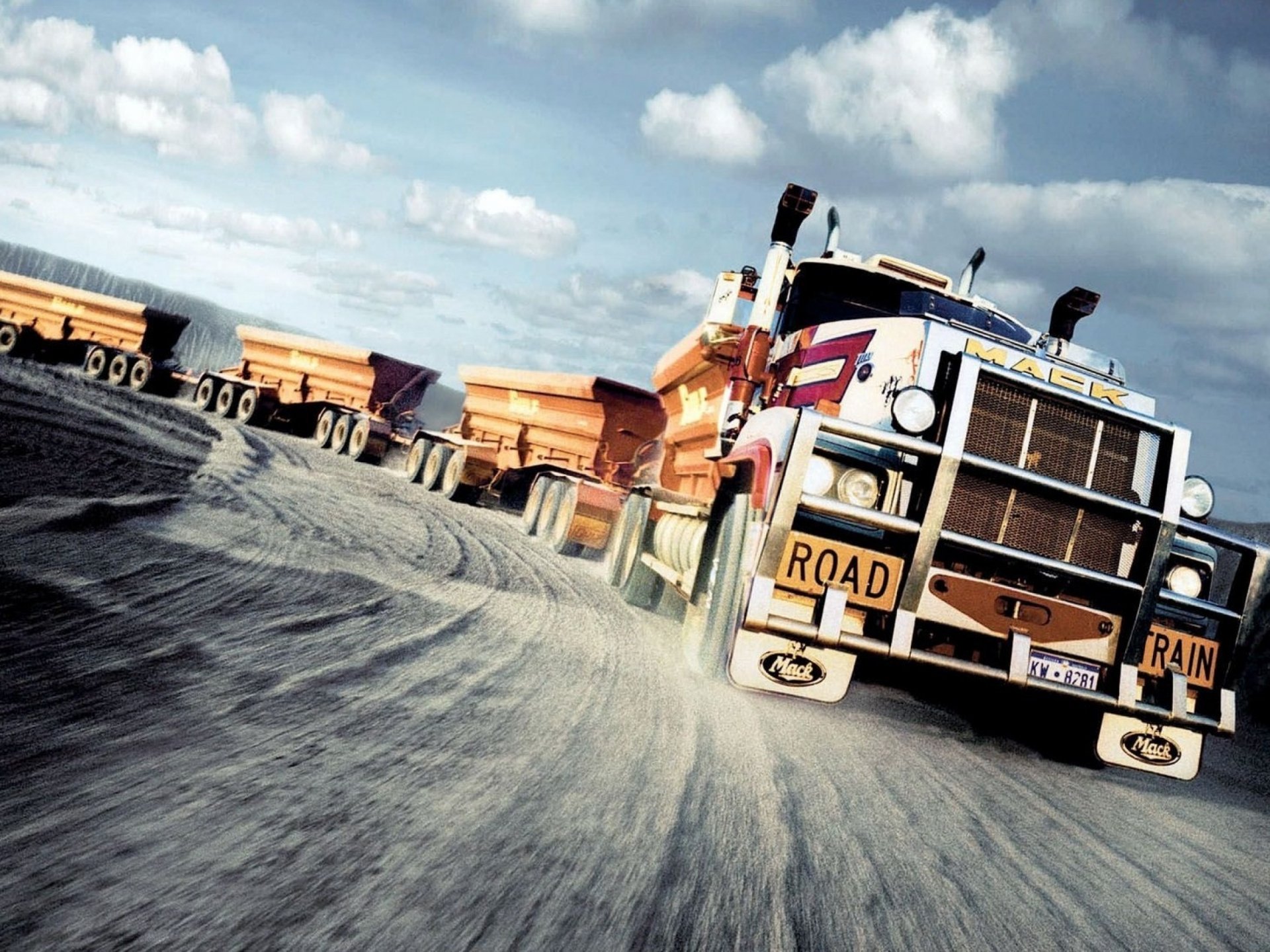 HD desktop wallpaper featuring a powerful Mack Trucks vehicle driving on a dirt road with a ROAD TRAIN sign under a cloudy sky.