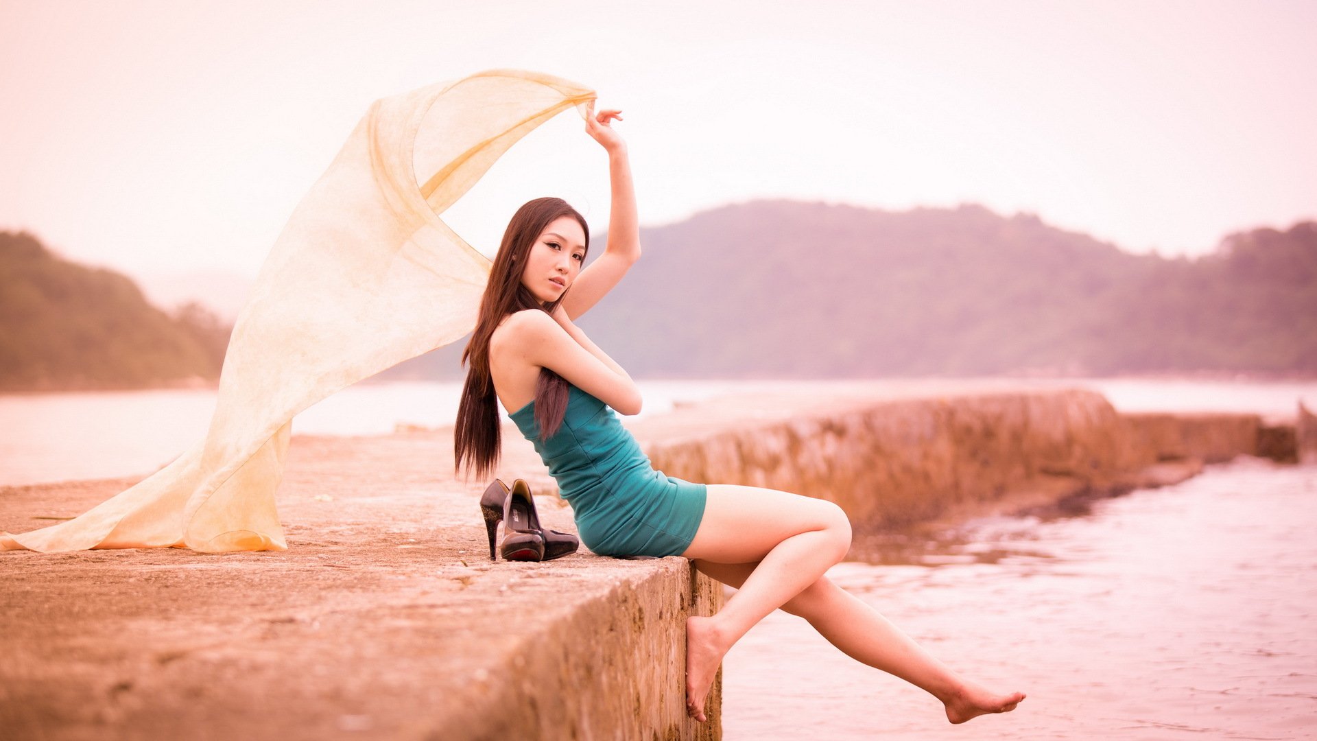 A woman sits gracefully on a ledge by the water, draped in a light scarf, exuding elegance against a serene landscape. This HD desktop wallpaper features soft hues and a tranquil atmosphere.