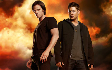 Two main characters from the TV show Supernatural stand confidently against a backdrop of fiery clouds, showcasing their striking presence in this HD desktop wallpaper.