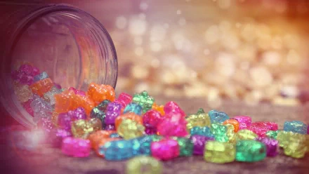 Colorful, shiny candy spills from a tipped jar, creating a vibrant display on a soft surface. This delightful image serves as an HD desktop wallpaper and background.