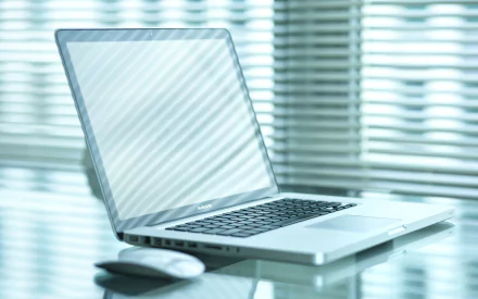 A sleek MacBook Pro sits elegantly on a glossy surface, accompanied by a mouse, against a backdrop of softly diffused light from window blinds. A modern tech-inspired desktop wallpaper.
