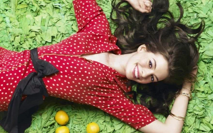 A vibrant HD desktop wallpaper featuring Anne Hathaway lying on green grass, wearing a red polka dot dress and smiling joyfully with oranges beside her.