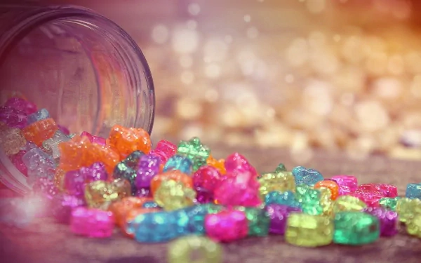Colorful, shiny candy spills from a tipped jar, creating a vibrant display on a soft surface. This delightful image serves as an HD desktop wallpaper and background.