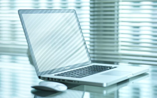 A sleek MacBook Pro sits elegantly on a glossy surface, accompanied by a mouse, against a backdrop of softly diffused light from window blinds. A modern tech-inspired desktop wallpaper.