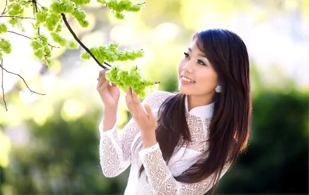 A woman is smiling while admiring green leaves in a natural outdoor setting. The background is blurred, emphasizing her white lace outfit and the vibrant greenery. The image is a HD desktop wallpaper.