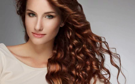 A striking redhead woman with long, wavy hair softly gazes at the viewer, set against a light backdrop, creating an elegant HD wallpaper for desktop backgrounds.