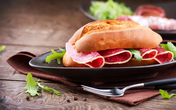 HD PC desktop wallpaper of a fresh sandwich with sliced salami and greens on a black plate, set on a rustic wooden table with a brown cloth and fork nearby.