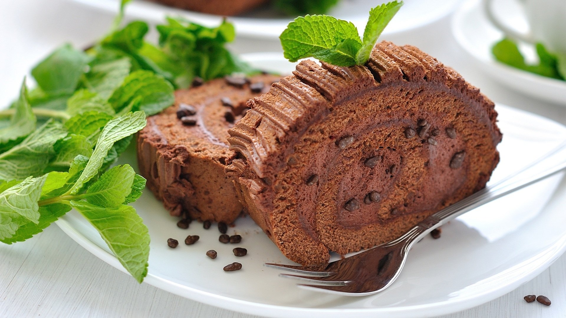 A delicious chocolate cake roll garnished with fresh mint leaves, surrounded by chocolate chips, creating an enticing food-themed HD desktop wallpaper.
