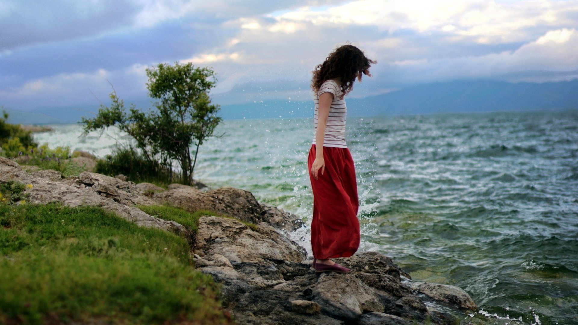A woman in a red skirt stands contemplatively by the rocky shoreline of a large body of water with distant mountains under a partly cloudy sky. This HD desktop wallpaper captures a reflective mood.