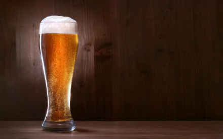 A crystal-clear glass of golden beer with frothy foam sits on a wooden surface, against a warm, dark background, creating an inviting food and drink atmosphere.