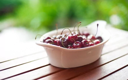 HD desktop wallpaper featuring a white bowl filled with fresh cherries on a wooden surface, set against a blurred green outdoor background.