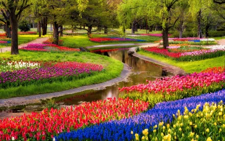 HD desktop wallpaper featuring a beautifully photographed park with colorful flower beds, meandering paths, and a tranquil stream surrounded by lush greenery and vibrant blossoms.