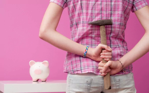 Person hiding a hammer behind their back beside a man-made piggy bank on a table, pink backdrop — 2K Quad HD PC desktop wallpaper and background.