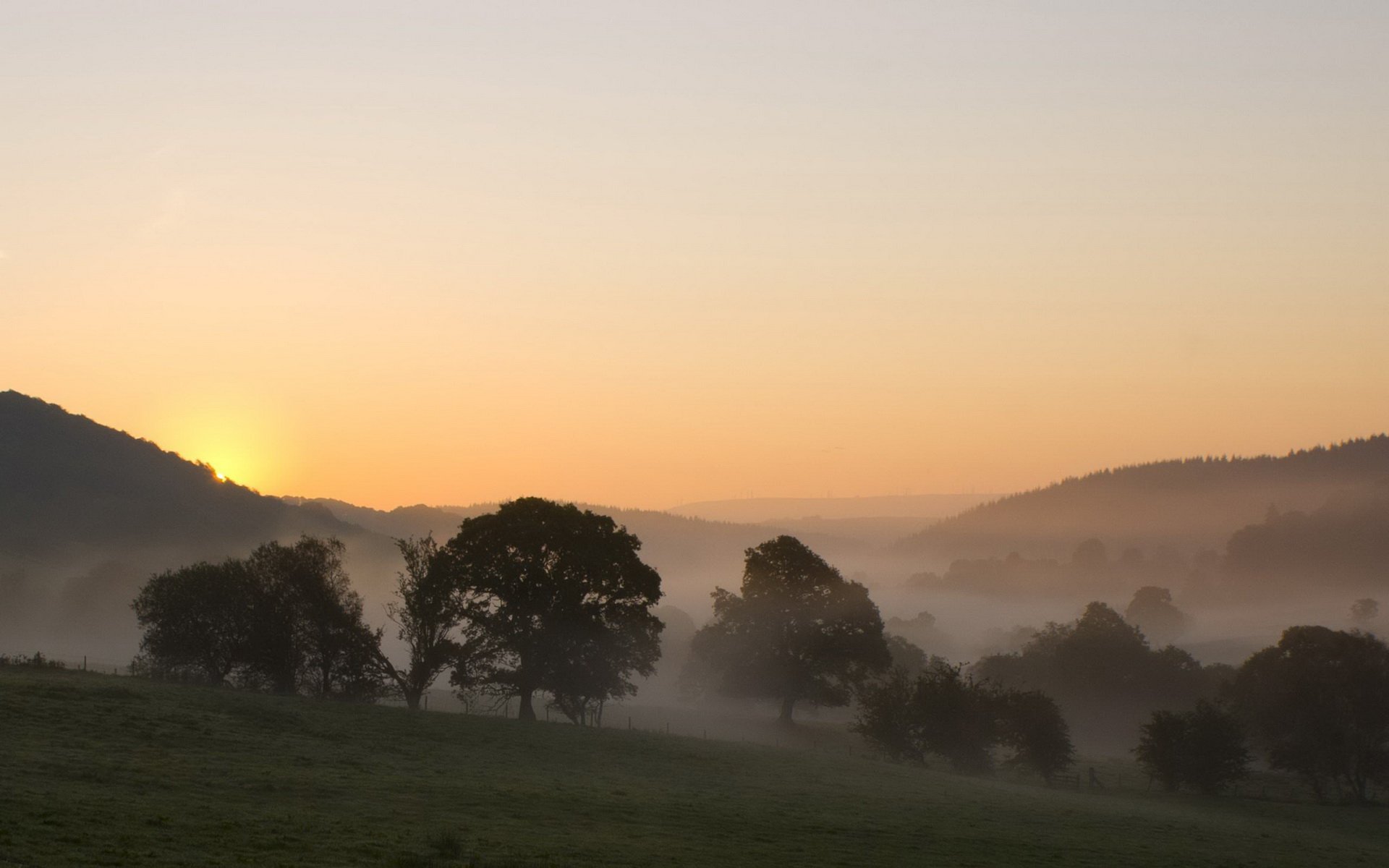 HD PC desktop wallpaper: foggy nature scene at sunrise, low mist rolling through a valley with silhouetted trees and distant hills.