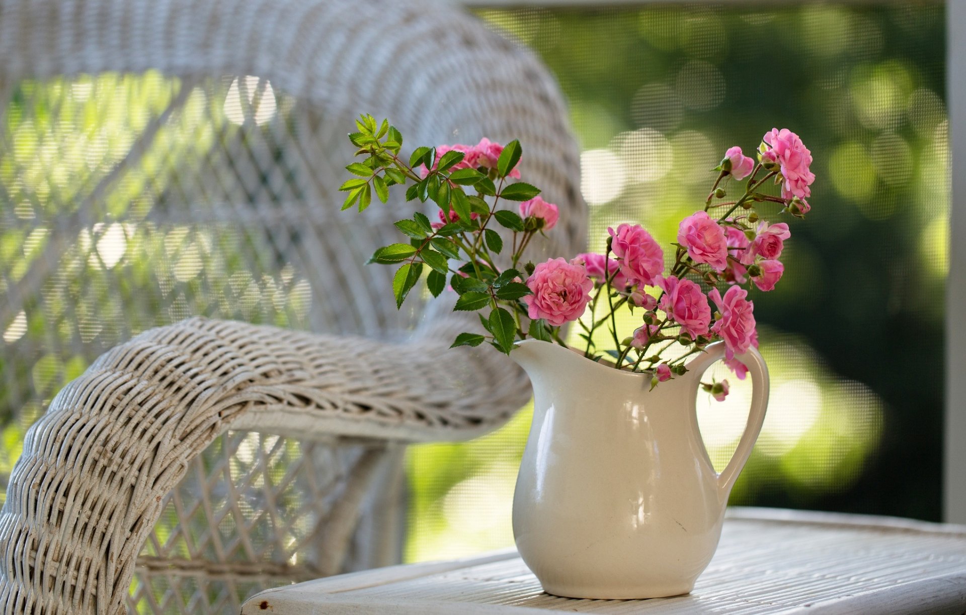 A serene HD desktop wallpaper featuring a white pitcher filled with vibrant pink flowers, set beside a wicker chair. The soft background enhances the floral arrangement's beauty.