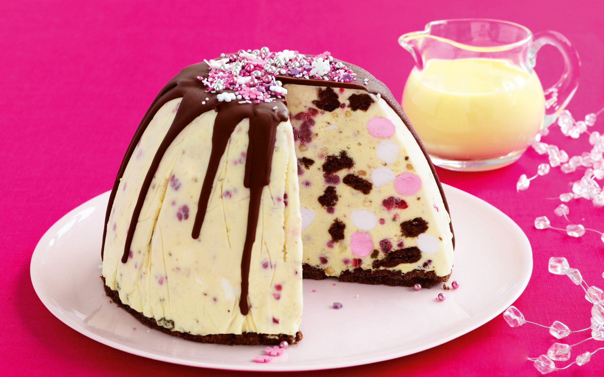 HD PC desktop wallpaper featuring a colorful cream cake with chocolate drizzle and sprinkles on a white plate against a vibrant pink background, accompanied by a glass pitcher.