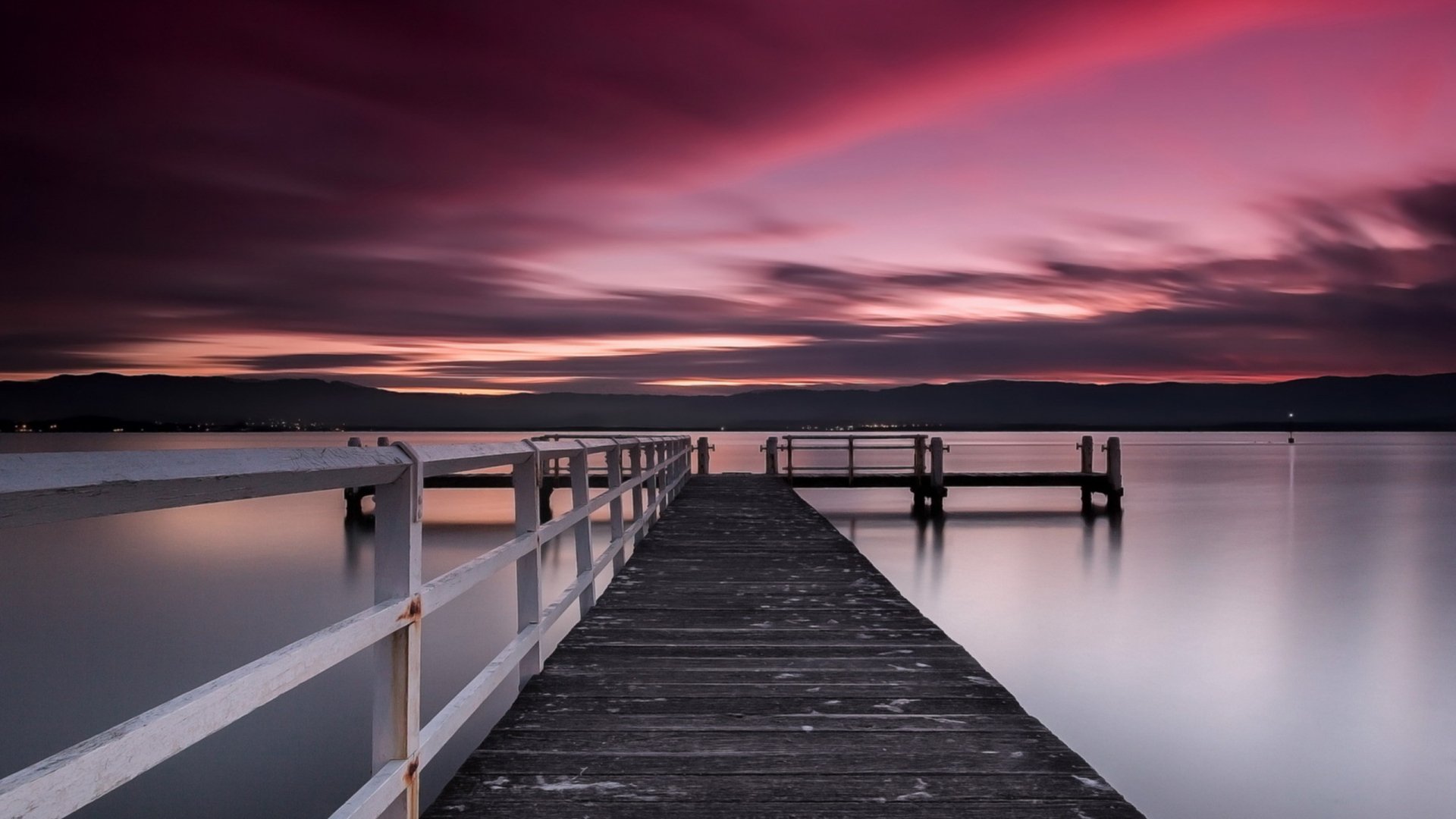 A serene pier extends into calm waters under a vibrant sunset, creating a tranquil scene. This HD wallpaper captures the beauty of man-made structures in nature.