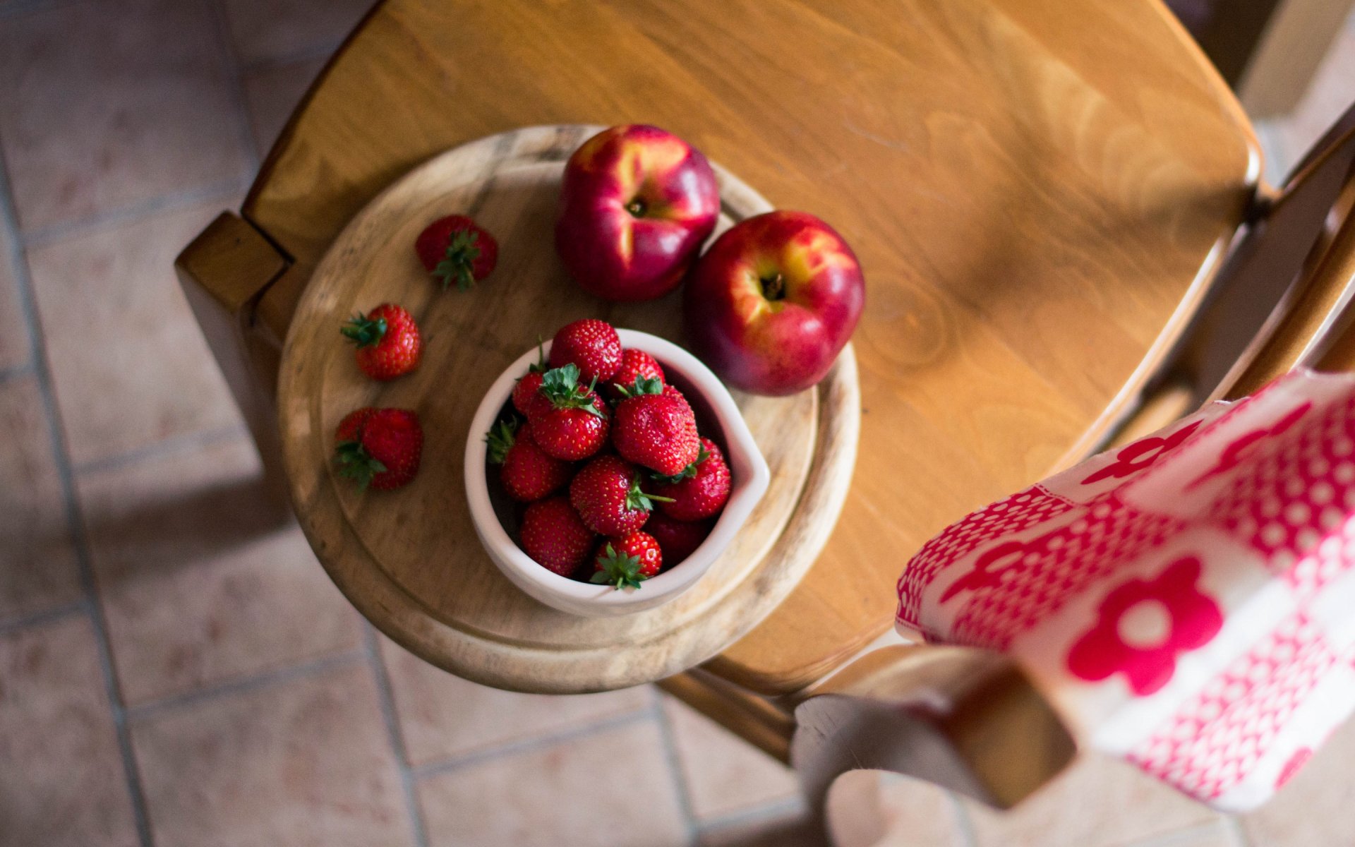 HD desktop wallpaper featuring fresh strawberries, two apples, and a few tomatoes arranged on a wooden stool with a red and white cloth draped over a nearby chair.