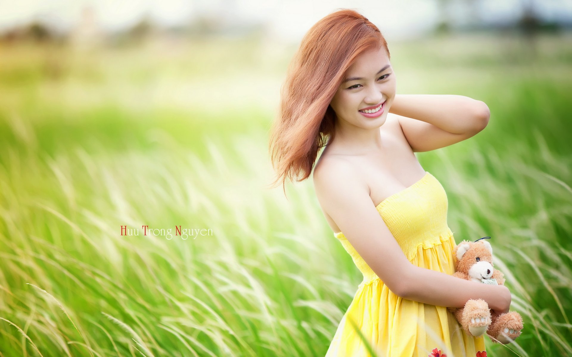 A smiling Asian woman in a yellow dress poses in a field of tall grass, holding a teddy bear. This vibrant HD image serves as an engaging desktop wallpaper.
